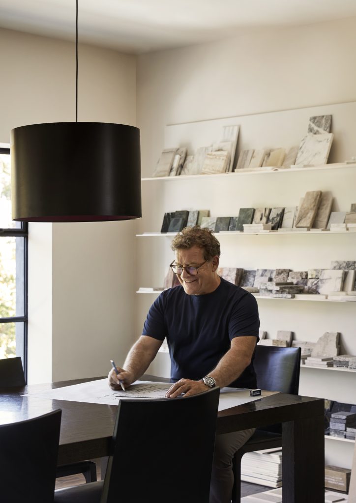 Rob Mills Architects. A middle aged man in a dark T shirt and glasses sits at a desk under a large lampshade drafting something on a big sheet of paper