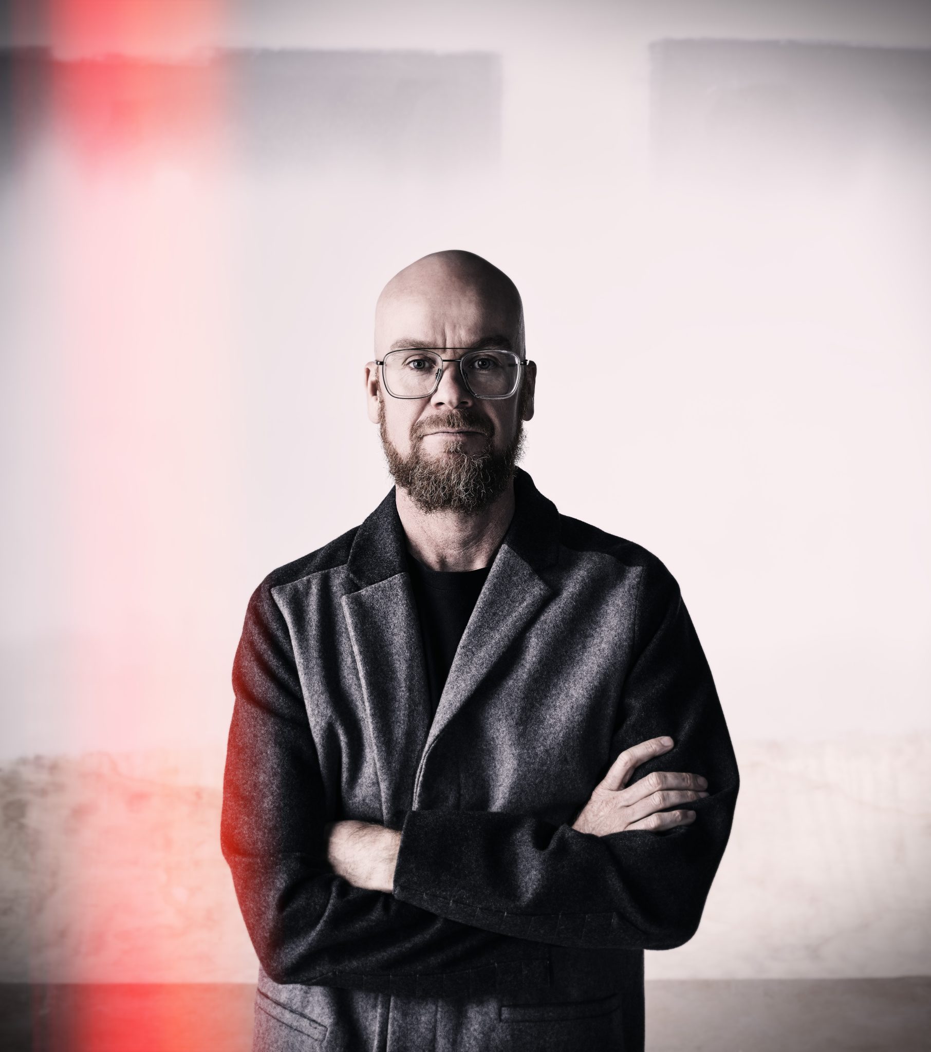 Ross Gardam. A bald headed, bearded man wearing glasses, stands with his arms folded looking at the camera. He is wearing a grey jacket with black lapels over a black T shirt. The image has a orangey stripe that fades in the centre up the left hand side.
