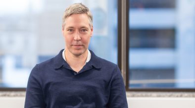 Simon Fleet sits at a desk looking at the camera and with clasped hands and wearing a blue jumper. He has blond hair streaked with grey and is looking at the camera with a slight smile.