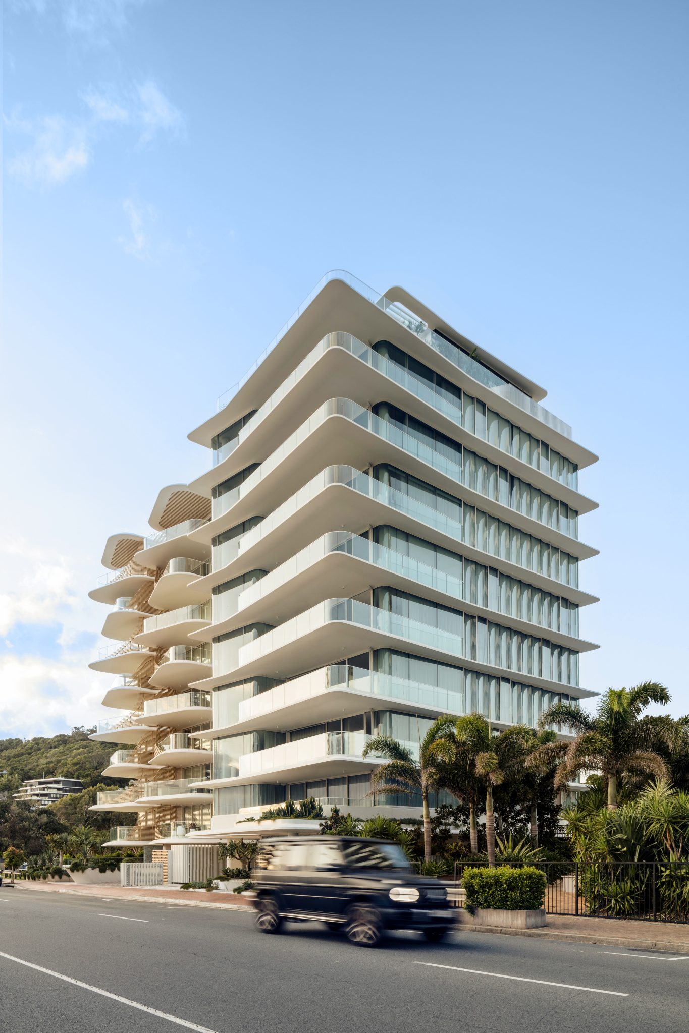 vertical city, image of a medium high rise block with prominent balconies and one behind. A black car is driving across the road in front.