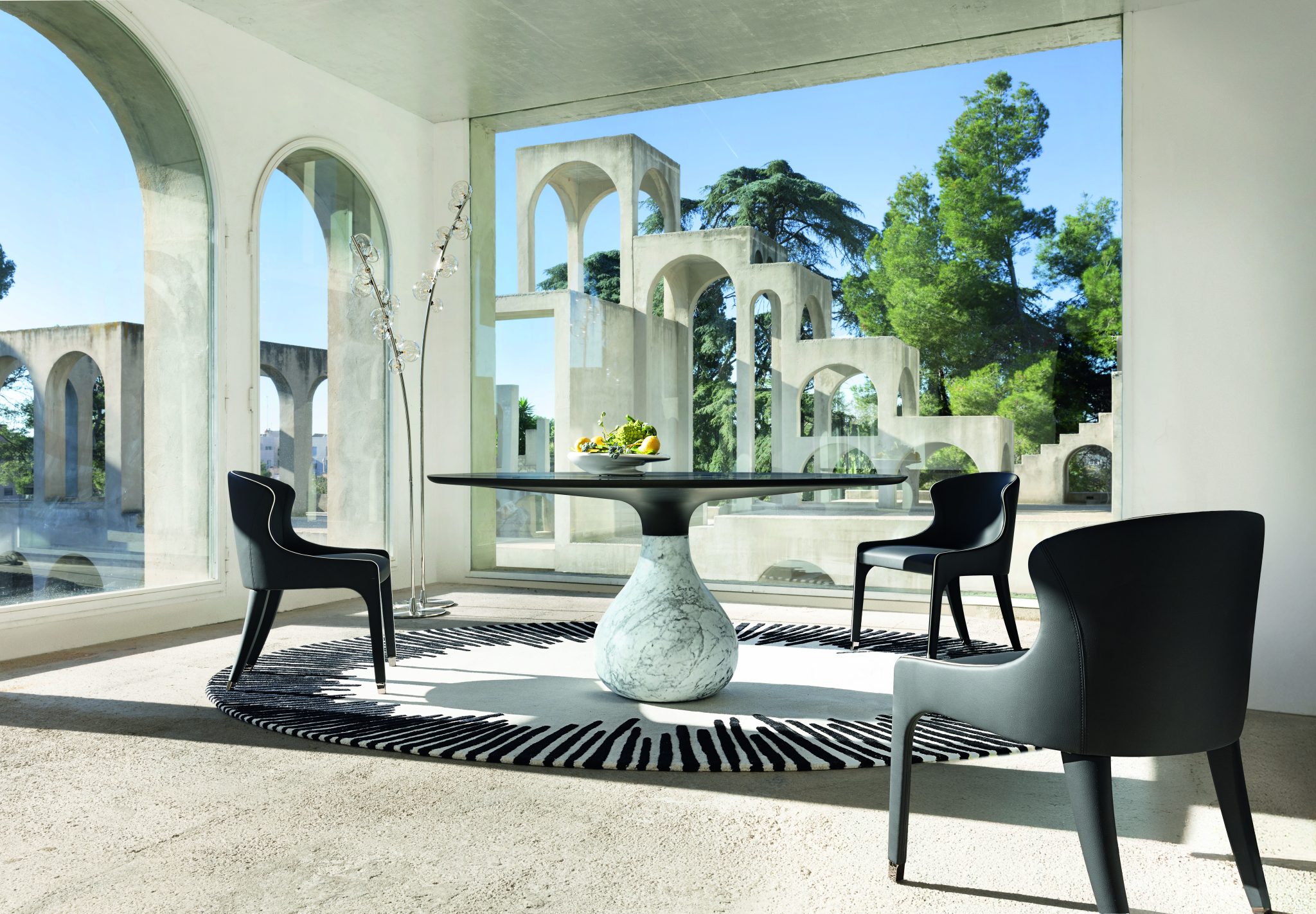 Aqua table – Roche Bobois, a black topped circular table with a bulbous marble base, surrounded by three black chairs, on a white rug with black stripes coming in from the edge part of the way, inside a geometric Escher like construction, with trees outside