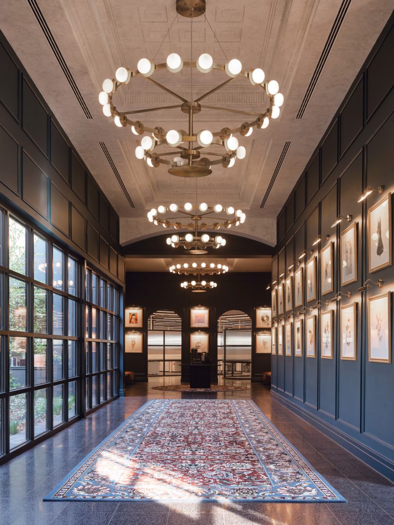 Designer Rugs. A grand hallway with several chandeliers reaching back into the shot, windows on the left, paintings hung in a row of two on the right and a long designer run traversing the space.