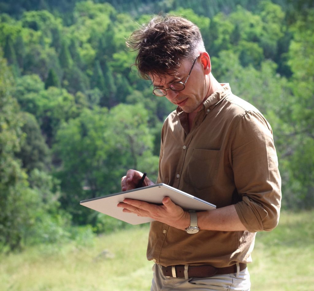 Jiri Lev - a bespectacled man in a brown shirt and light trousers is holding a large tablet and using a stylo with it