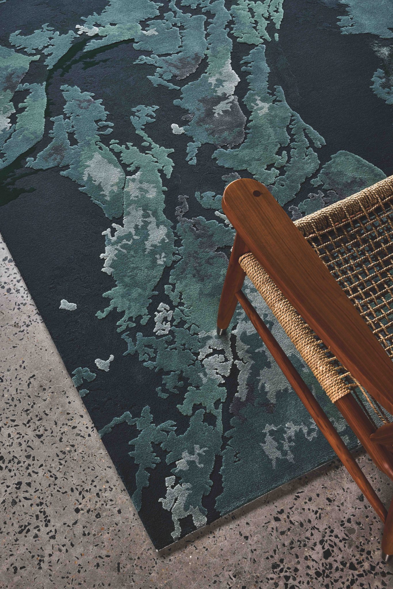 Designer Rugs. Detail of a large rectangular rug in different shades of green, with patterns that look like Scandinavia but aren't. The corner of a wooden chair with a woven seat is in the bottom right hand corner of the frame.