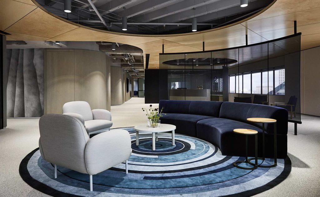 Designer Rugs. A hotel lobby space with grey armchairs, a black bench seat and a small table, all on top of a circular blue and white designer rug.