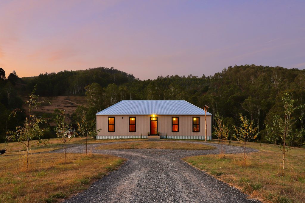 Jiri Lev, a simple wooden house seen at dusk, with woods and a hill behind.