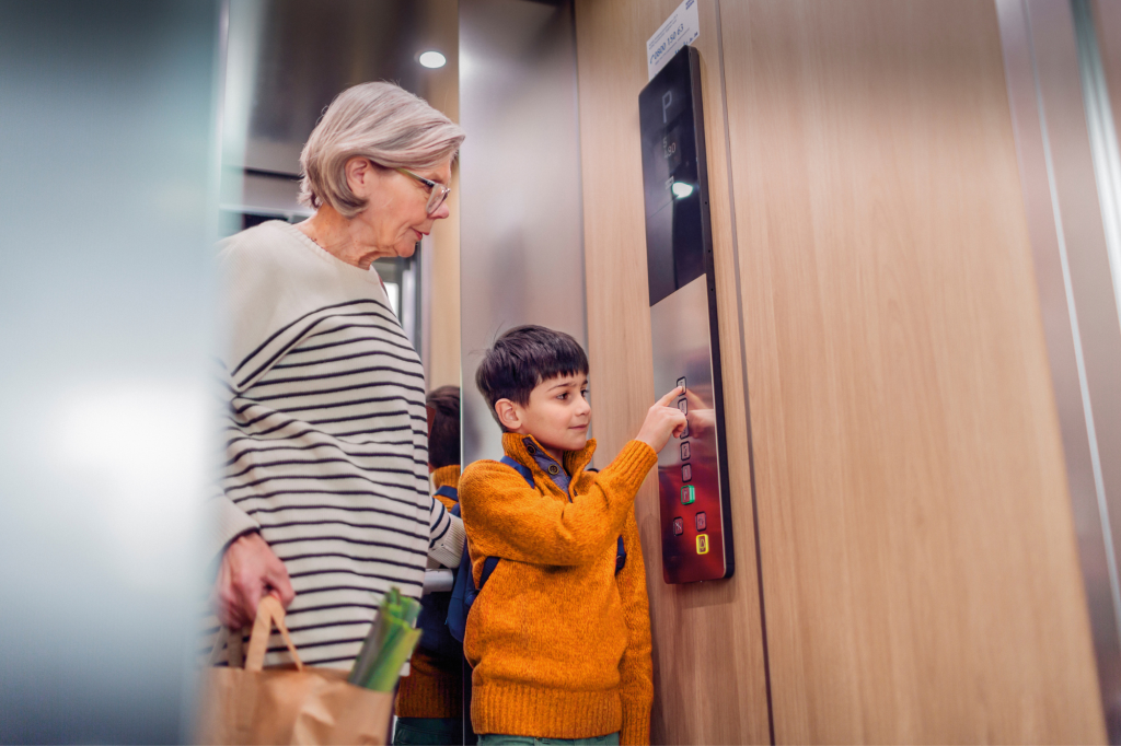 rethinking density. An older gray haired woman in a horizontally striped top and a young dark haired boy in an orange jacket are in a lift. She is carrying shopping, he is pushing the buttons.