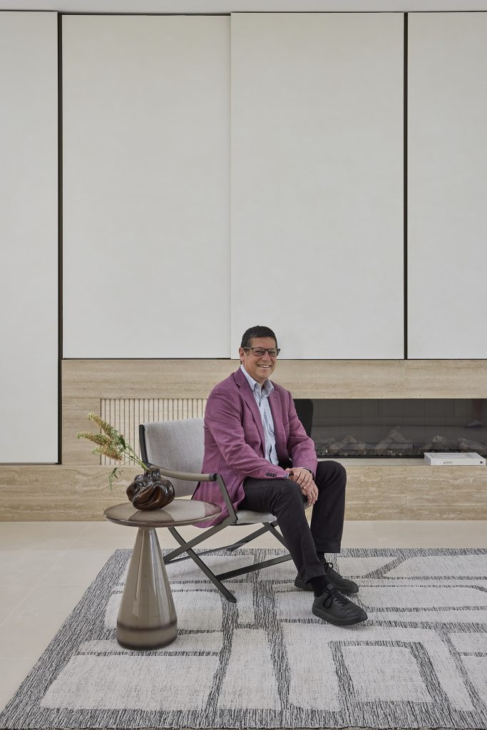 Designer Rugs. A man in a pinkish jacket and black trousers sits in a grey chair in the middle of the frame, next to a small side table with a minimalistic floral display. Underneath him is a grey and white designer rug with geometric patterns.